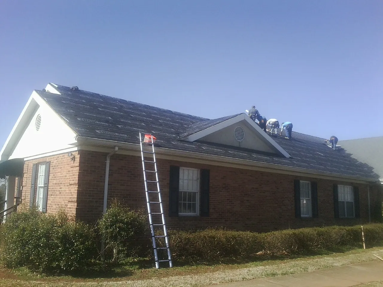Crew tearing shingles off a brick ranch roof with a ladder set — Emergency Roof Repair work by our crew in West Glacier
