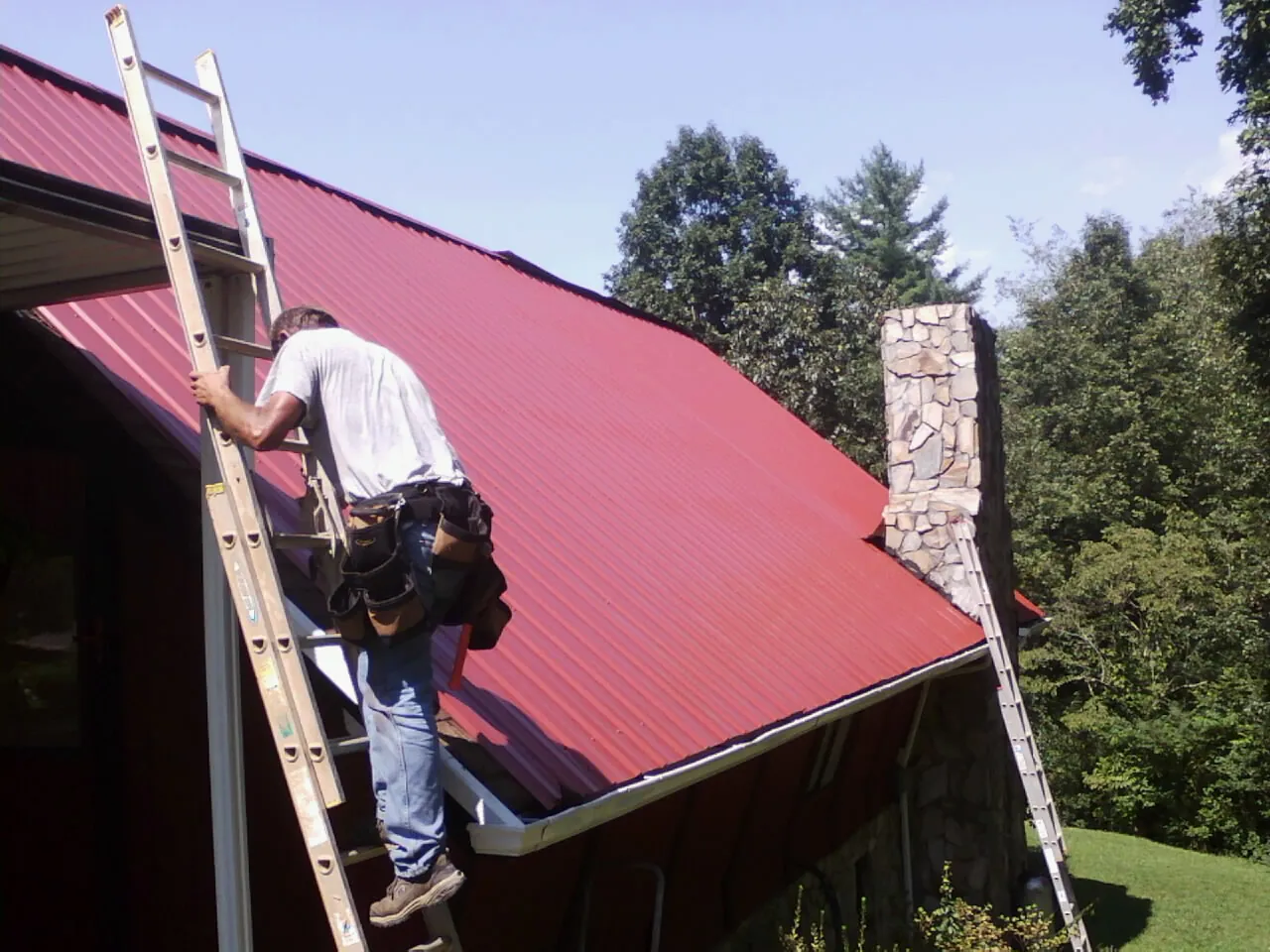 Roofer on a ladder beside a red metal roof and stone chimney — Soffit & Fascia Repair by Roofing Brothers in West Glacier